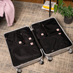 An open silver hard-shell suitcase on a patterned rug, showing black hae hae packing cubes and organizer pockets inside. A pink knit blanket, books, and a potted plant are visible in the room setting.