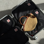An open black silver rolling suitcase shown from above, packed black hae hae compression packing cubes, a woven straw bag, sunglasses, and a passport, on a gray patterned rug.