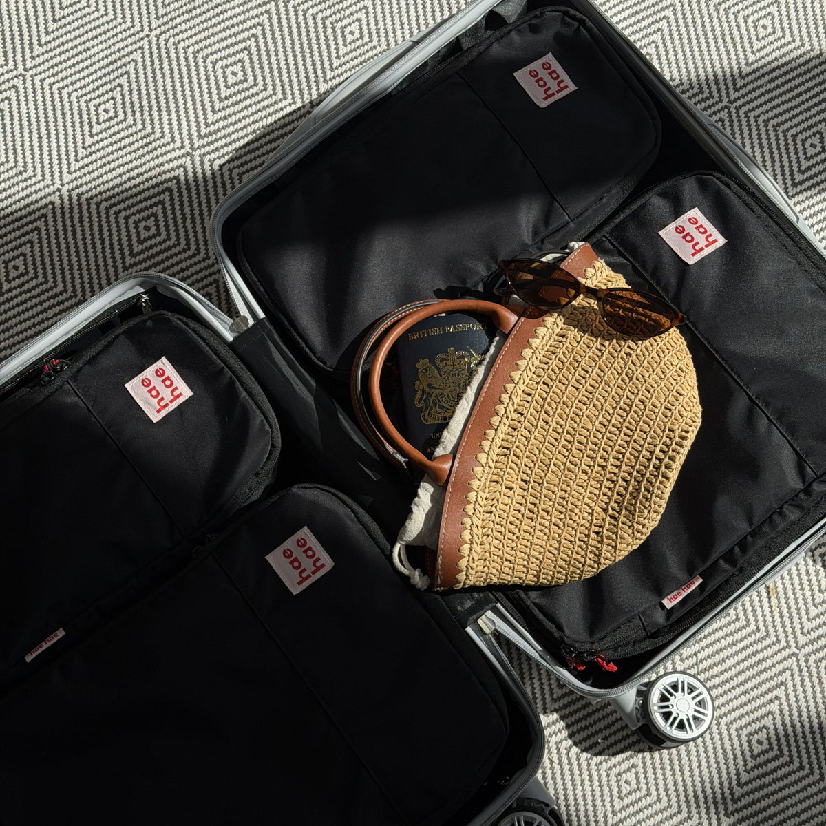 An open black silver rolling suitcase shown from above, packed black hae hae compression packing cubes, a woven straw bag, sunglasses, and a passport, on a gray patterned rug.